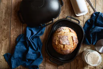 A baked loaf sitting in the cast iron base of the Challenger Bread Pan, with the lid set to one side